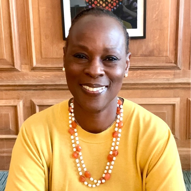 The image features an individual smiling at the camera. They have a warm and inviting expression, with short-cropped hair and clear skin. They are wearing a bright yellow, round-neck top, complemented by a double-stranded necklace adorned with larger beads that are white and a translucent orange. The necklace adds a vibrant touch to the attire. The individual appears to be in a room with wooden furnishings and a painting with orange tones visible in the background, which harmonizes with the necklace's colors. The overall feel of the image is friendly and approachable.