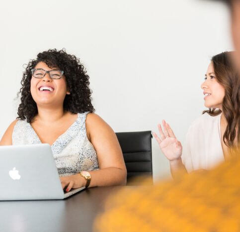 Two women are engaged in a lively conversation in a modern office setting. The woman on the left, with curly hair and glasses, is laughing joyfully while working on her laptop. She is dressed in a light-colored, sleeveless top. The woman on the right, partially visible, appears to be speaking animatedly, wearing a light blouse. The atmosphere is friendly and collaborative, suggesting a positive and engaging work environment. A blurred figure in the foreground adds depth to the image.