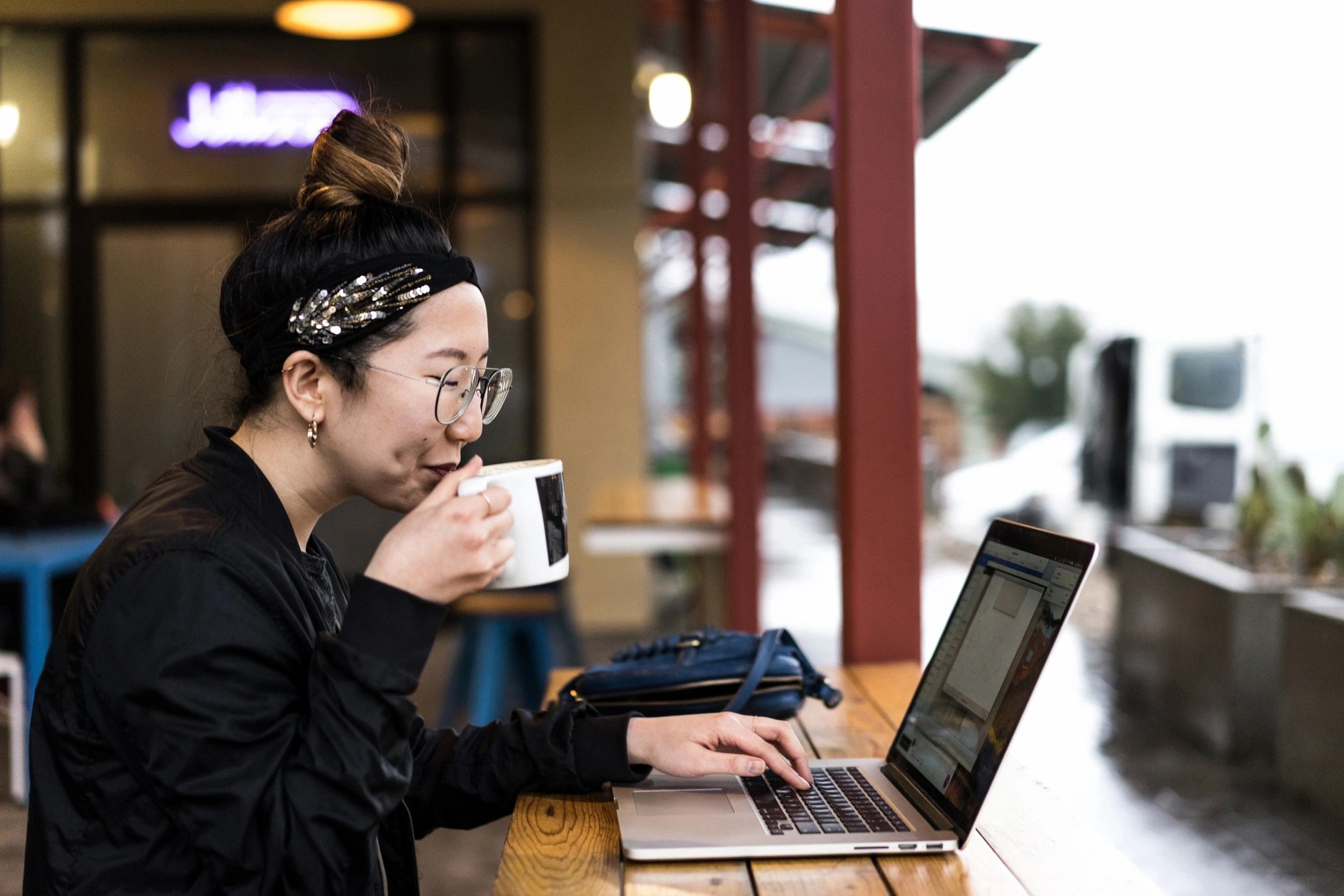 O the Leading Change in Healthcare page, this is an image of a woman wearing glasses and a black headband with decorative details is sitting at a wooden table in an outdoor café. She is sipping from a white mug and working on a laptop. The background shows a blurred view of the café's interior and a rainy scene outside.