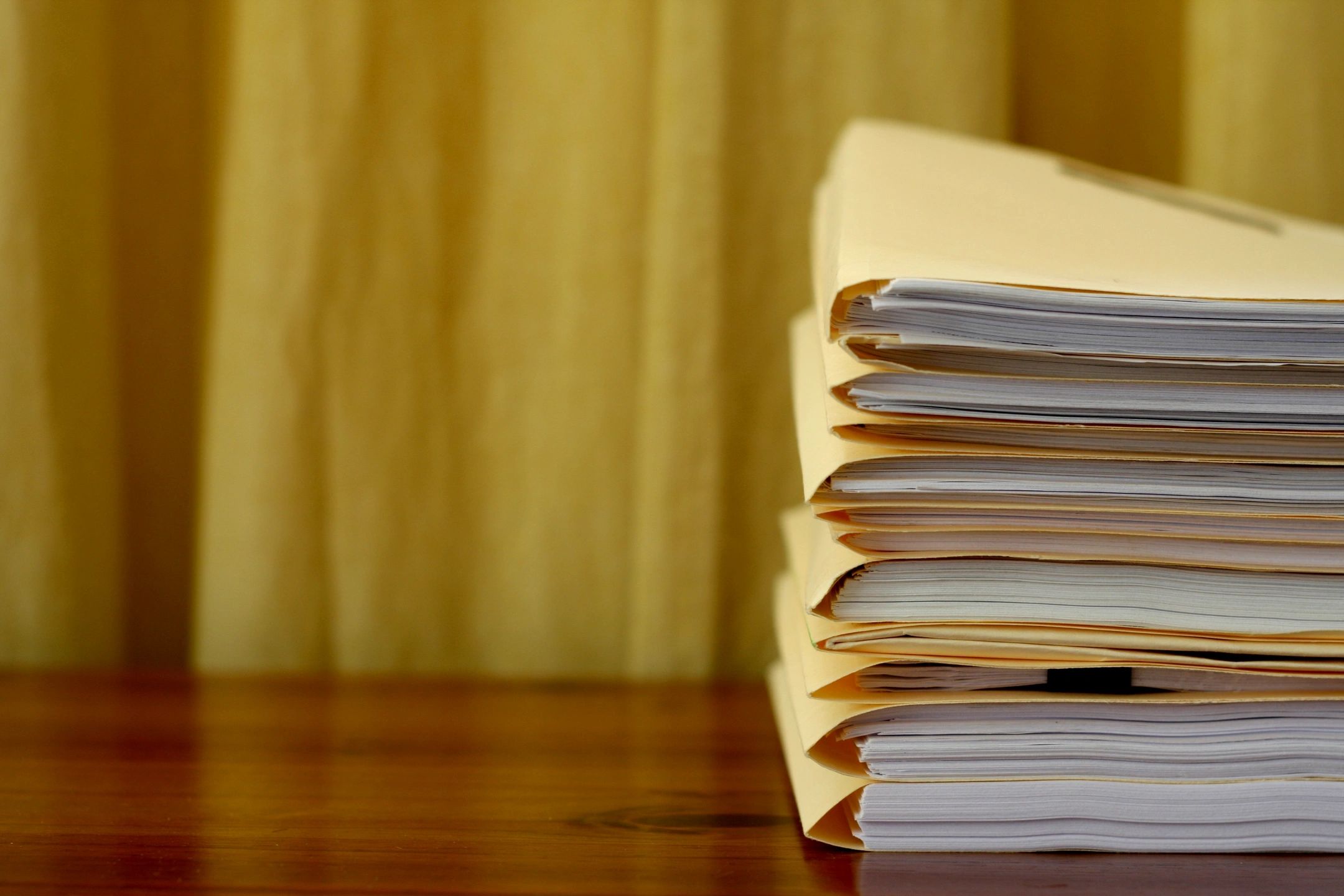 A stack of manila folders filled with papers, neatly organized and placed on a wooden surface against a soft yellow curtain backdrop. The image conveys a sense of organization and thorough documentation.