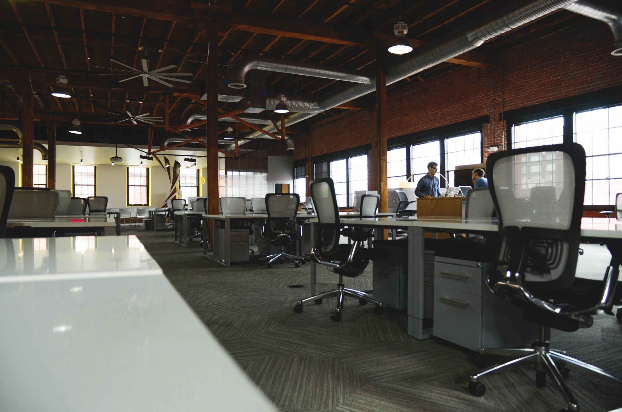 An open-plan office with modern, industrial design elements, featuring exposed brick walls, large windows, and a wooden ceiling with exposed beams. The workspace is equipped with ergonomic office chairs and desks, with a few people conversing in the background, suggesting a collaborative and productive environment.