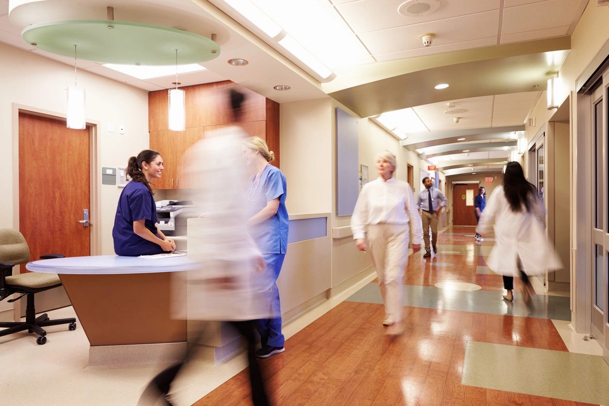 Healthcare professionals and staff moving through a busy hospital corridor, with some engaging at a nurse's station, highlighting the dynamic and fast-paced environment of a healthcare facility.