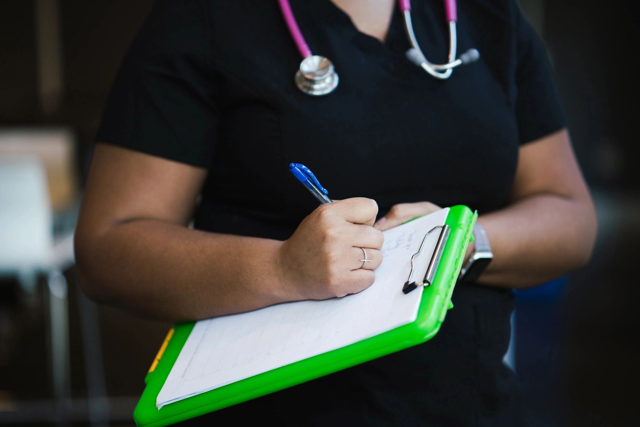 Healthcare Trends. Here is a description for the second image you uploaded: A healthcare professional, wearing a black uniform and a stethoscope around their neck, is seen writing on a clipboard with a bright green border. The person's face is not visible, focusing attention on the act of writing and the stethoscope. The blue pen in their hand contrasts with the green clipboard, suggesting a clinical setting. The background is slightly blurred, ensuring that the primary focus remains on the healthcare professional's hands and the clipboard.