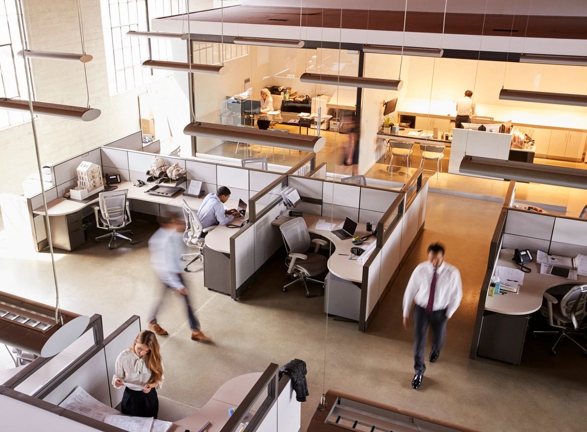 An open office space with modern cubicles and workstations, where employees are engaged in various tasks. Some individuals are walking through the office, while others are seated at their desks working on computers. The setting reflects a busy and collaborative work environment with natural light streaming through large windows.