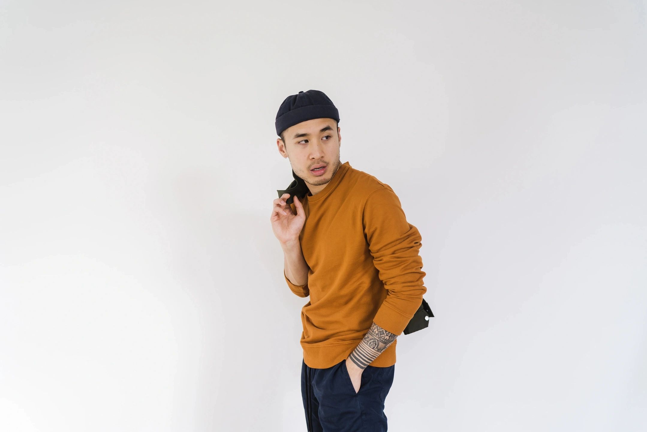 A young man in an orange sweater and blue pants poses against a plain white background. He is wearing a black beanie and has a tattoo on his forearm. His right hand is raised near his face, and he gazes to his left with a thoughtful expression