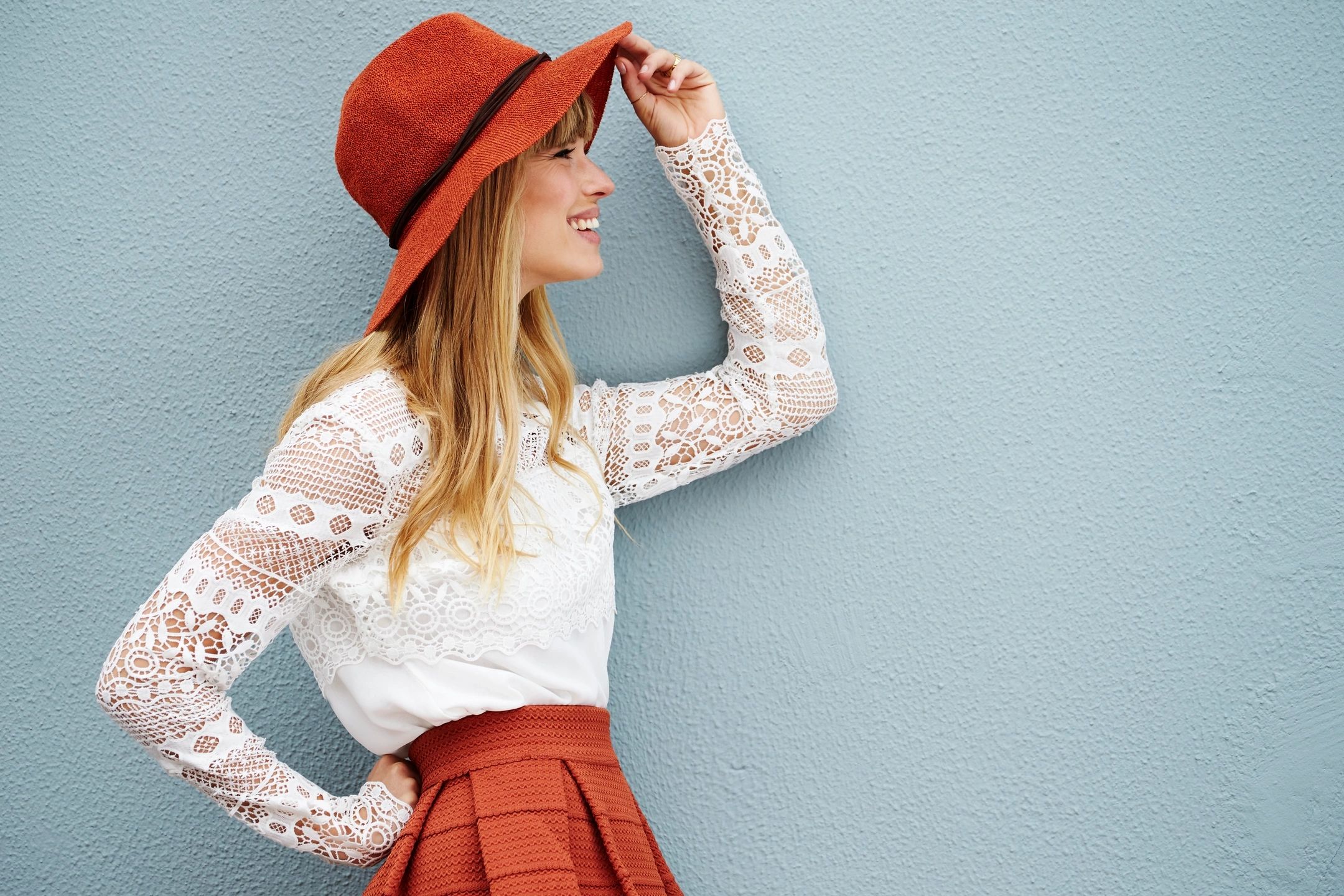 A young woman wearing a stylish red hat and a white lace top, smiling and posing against a light blue wall. She looks relaxed and confident, exuding a sense of modern fashion and individuality.