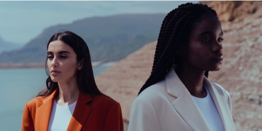 Two women are pictured outdoors against a backdrop of mountains and water. The woman on the left has long dark hair, wears an orange blazer, and looks serene, gazing into the distance. The woman on the right has long braids, wears a white blazer, and stands with a composed expression, facing slightly away from the camera. The scene conveys a sense of confidence, individuality, and connection with nature.