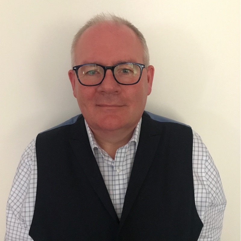 On the Meet the Team page, this is a middle-aged man with short, light hair and glasses is pictured against a plain white background. He is wearing a checked shirt and a dark-coloured vest. His expression is neutral and professional, presenting a composed and approachable demeanour.