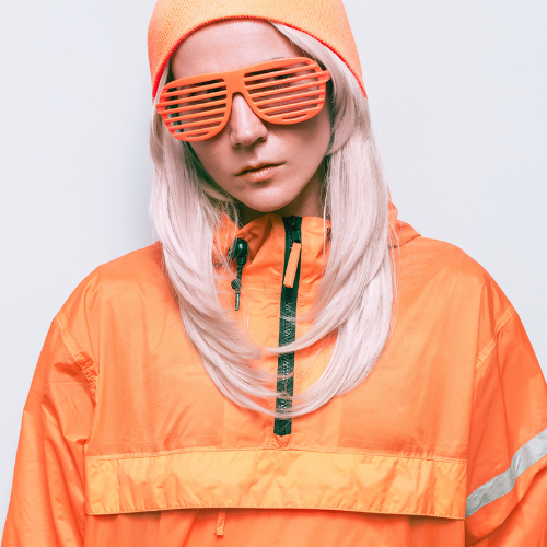 This image shows a person dressed in bright orange attire, including a beanie, jacket, and stylized shutter shades, with long, light-colored hair. The overall look conveys a futuristic or high-contrast fashion style. Let me know if you'd like this description expanded or need edits for the image.