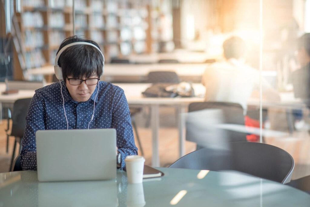 A focused individual wearing headphones and working on a laptop in a bright, modern library or co-working space, highlighting the role of digital learning and remote work in creating social impact through accessible education and collaboration.