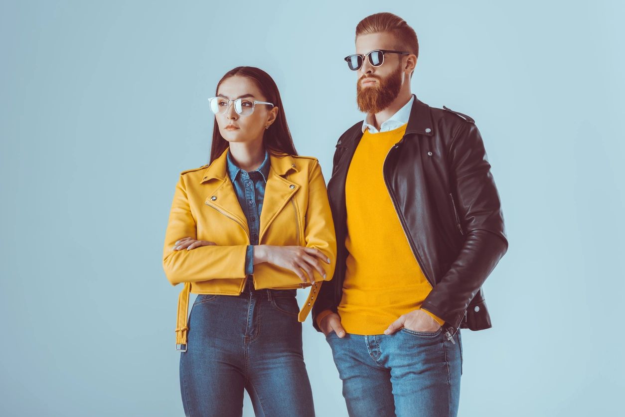 Two stylish individuals stand confidently against a light blue background. The woman wears a bright yellow leather jacket over a denim shirt, paired with blue jeans, while the man sports a black leather jacket, a vibrant yellow jumper, and blue jeans. Both wear sunglasses and pose with a cool, detached attitude, embodying confidence and individuality. This image aligns with the theme of tech transformations and change management, symbolising adaptability and modern style.